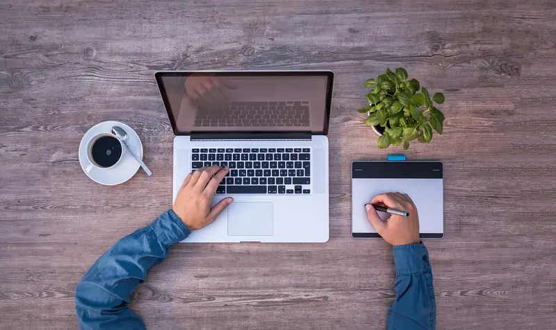 Hands use a laptop and a drawing tablet on a wooden table with coffee and a small plant, conveying a productive workspace.