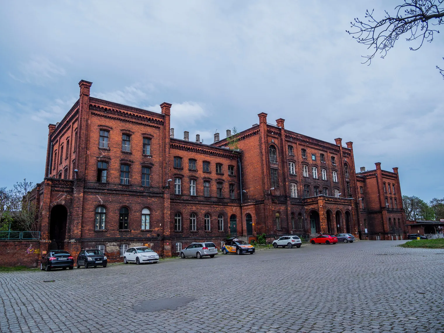 Historic red brick building with parked cars on a cobblestone courtyard under a cloudy sky.