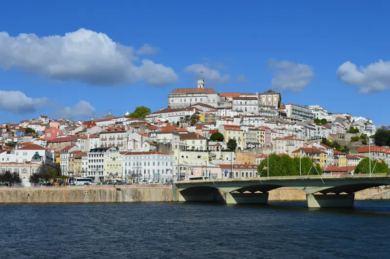 Hilltop city of white and pastel buildings above a river, with a bridge in front under a bright blue sky and clouds.