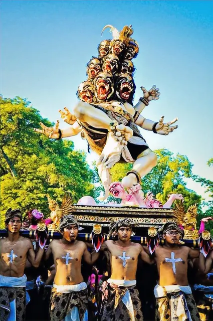 Men in traditional attire carry a large sculpture with multiple heads under a blue sky, creating a festive and vibrant atmosphere.
