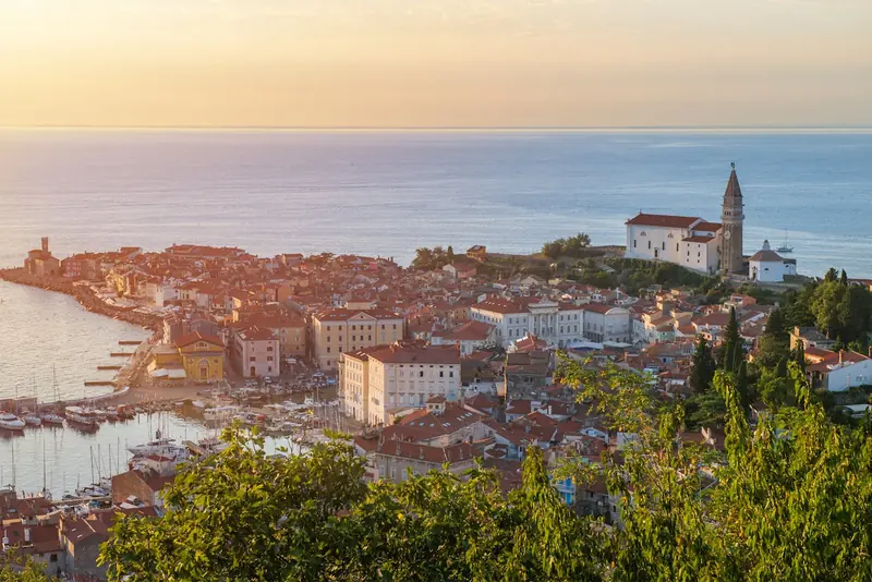 Scenic view of Piran, Slovenia, featuring red-roofed buildings, a church, marina with boats, and lush greenery in the foreground.