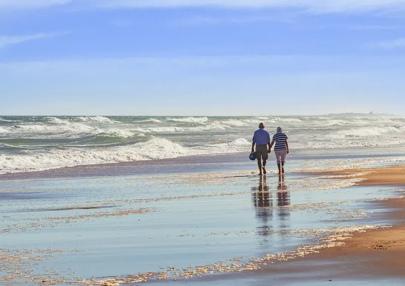 A couple walks hand in hand along the shoreline on a sunny day.