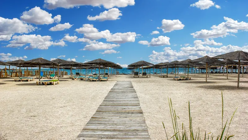 A wooden walkway leads to a sandy beach. Thatched umbrellas and sunbeds are scattered on the beach with the sea and blue sky in the background.