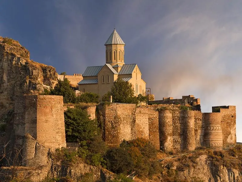 Narikala Fortress stands majestically atop a hill in Tbilisi, Georgia, with its ancient stone walls and central church creating a striking silhouette against the evening sky.