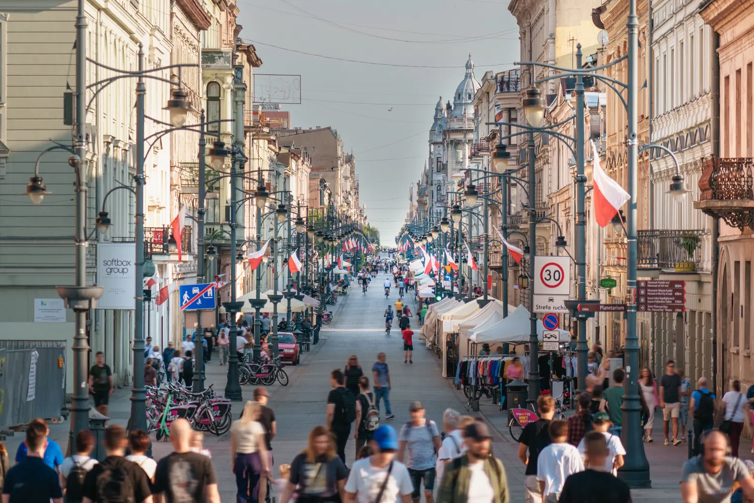 Busy pedestrian street lined with historic buildings, red-and-white flags, cyclists, and shoppers under a clear sky.