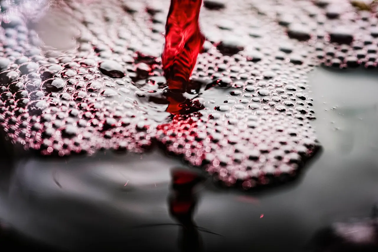 Red liquid pouring, creating bubbles on a dark surface. Close-up captures the dynamic, lively texture and rich color shades.
