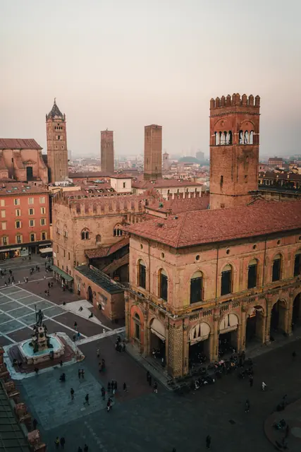 Aerial view of a historic brick square with towers, a fountain, and scattered pedestrians under a hazy sky.