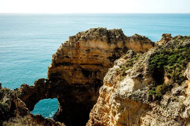 Rocky seaside cliffs with a natural arch above calm blue ocean under a clear sky, sunlit and serene.