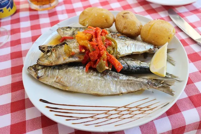 Grilled sardines with peppers and potatoes on a white plate, garnished with lemon and sauce, set on a red checkered tablecloth. The Sardine festival.