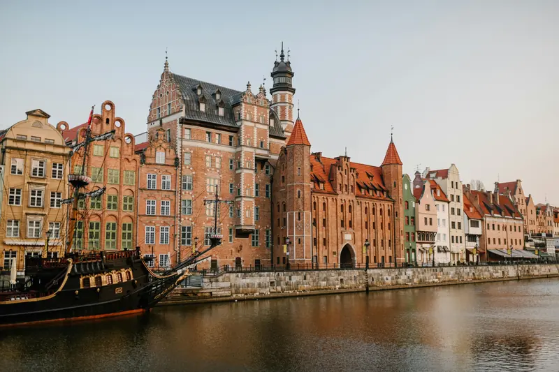 Historic red brick buildings and a replica ship along the Motława River waterfront in Gdańsk, Poland on a sunny day