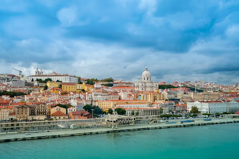 Colorful cityscape of Lisbon with pastel buildings and a large dome under a cloudy blue sky. A waterfront and mountains are visible in the distance.