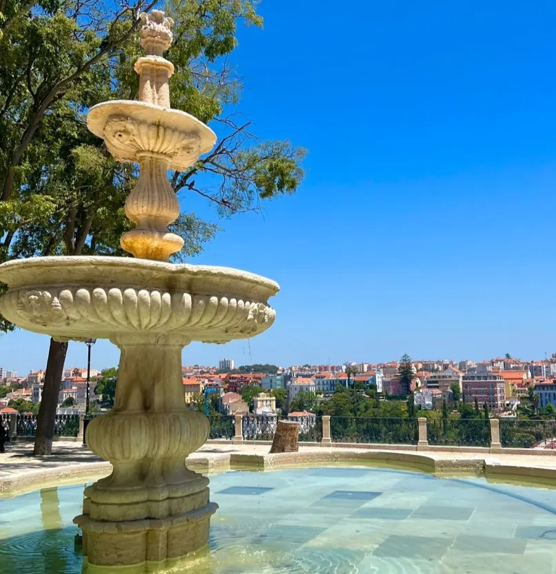 Stone fountain in a park overlooking a city skyline. Clear blue sky, trees, and colorful buildings in the background. Peaceful atmosphere.