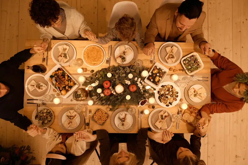 Family holding hands around a candlelit dinner table with festive dishes and greenery.