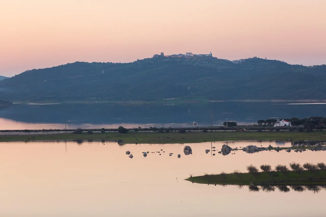 Serene sunset over a lake with green hills, reflecting pink skies. A distant village rests atop the hill, evoking a peaceful mood.