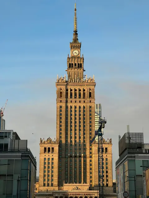 Tall historic building with a clock tower in the center, bathed in golden sunlight. Crane and modern buildings on either side. Clear blue sky.