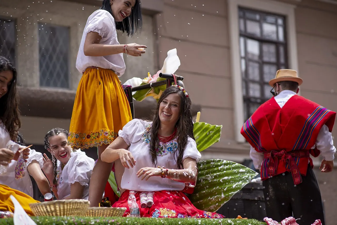 Women in colorful skirts celebrate with foam and laughter on a float in a festive street setting. A man in a red poncho is nearby.