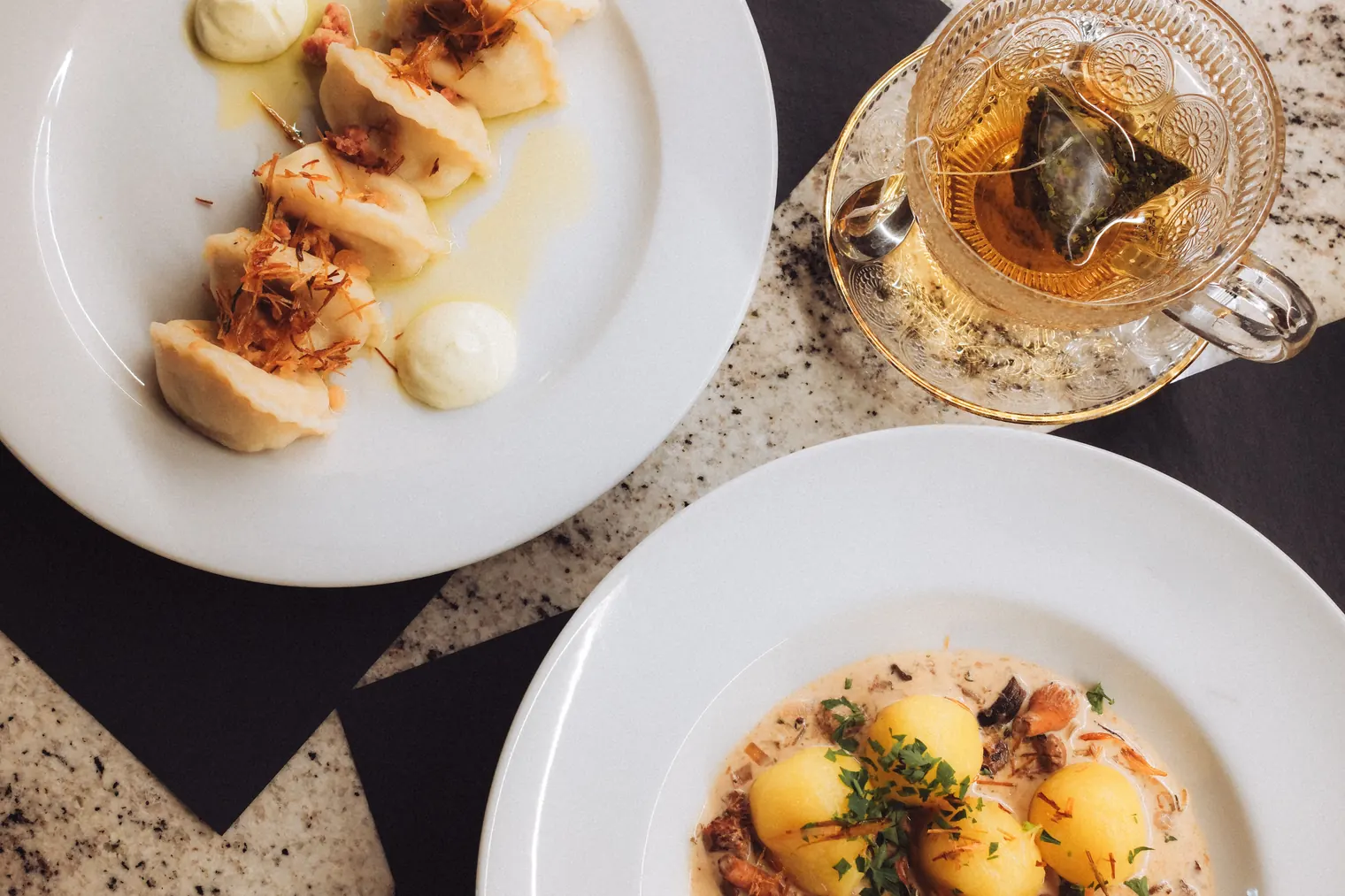 Overhead view of gourmet plated dumplings and a potato dish beside amber tea in a glass cup on a speckled table
