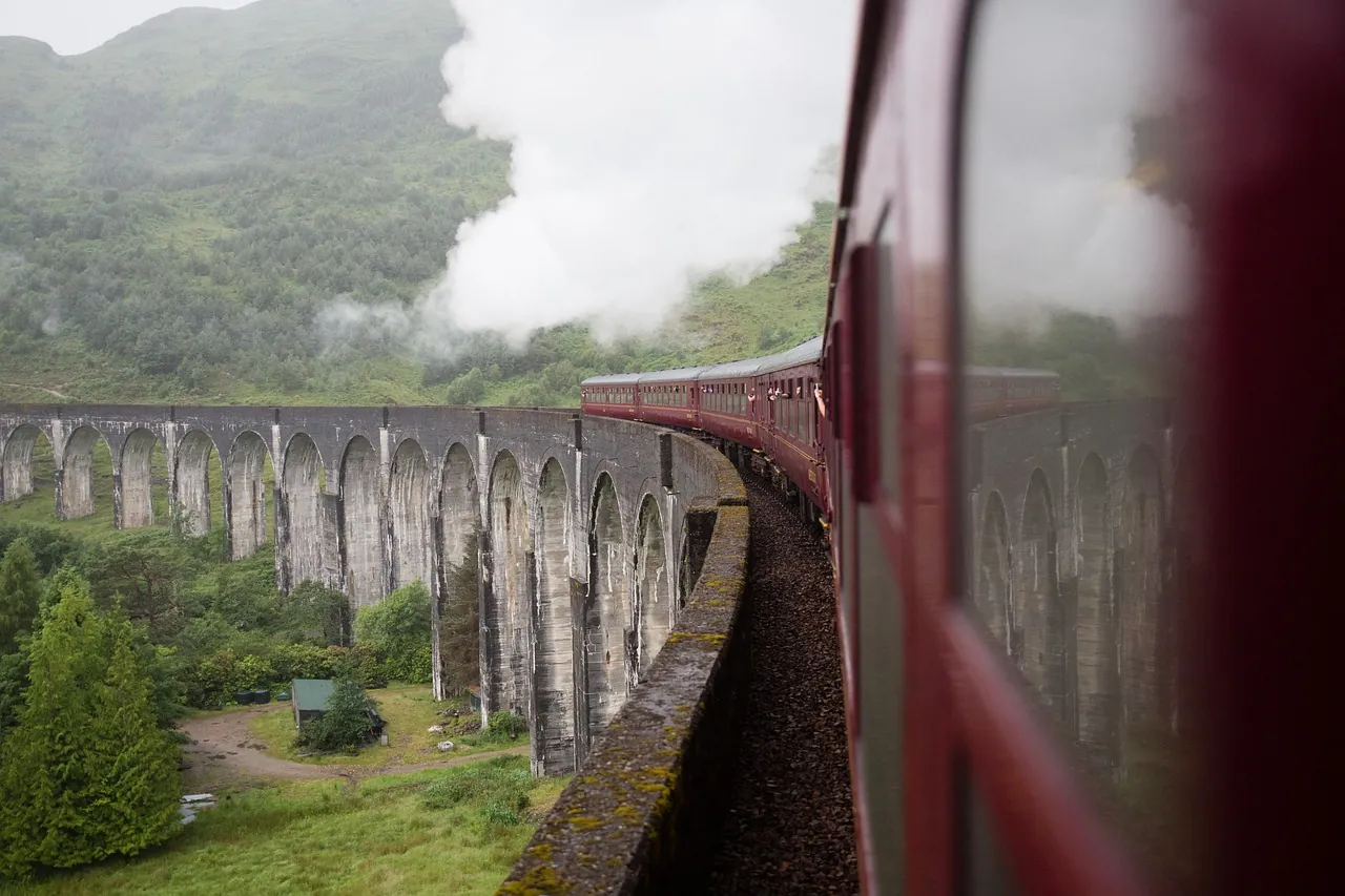 Red train crossing a stone viaduct in lush green hills, releasing steam. Overcast sky adds a moody atmosphere.