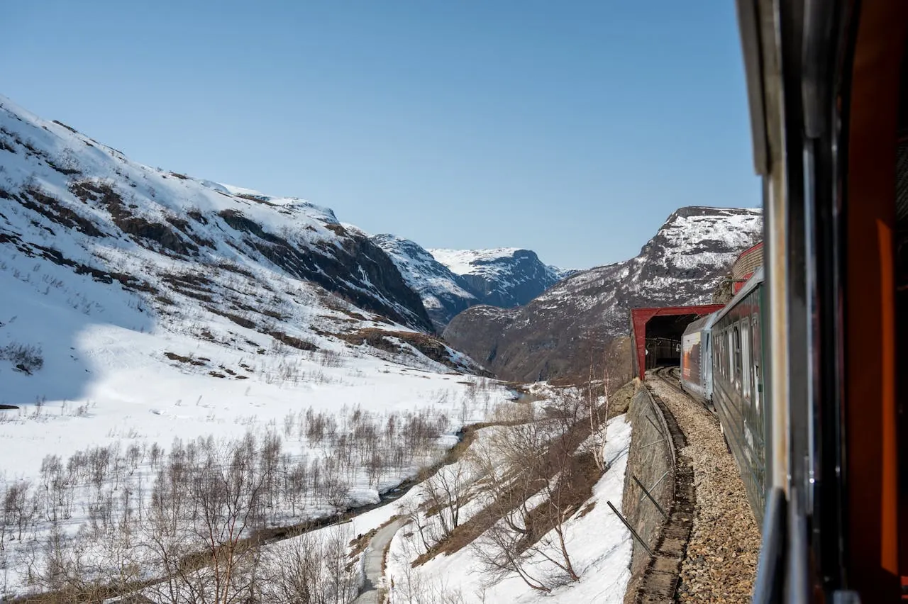 Train travels through snowy mountains under clear blue sky, surrounded by rocky slopes and leafless trees, evoking a serene winter scene.
