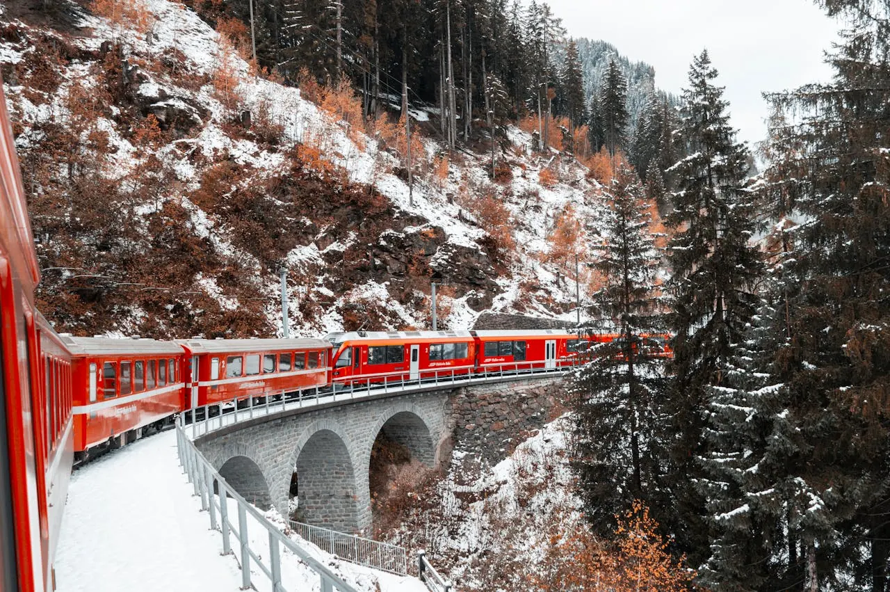 Red train crossing a snowy, arched stone bridge in a forested mountain landscape. Brown trees with snow create a serene winter scene.