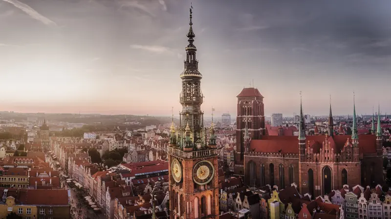 Aerial sunset view of a historic city with a tall clock tower and red-roofed cathedral, warm light over dense rooftops.