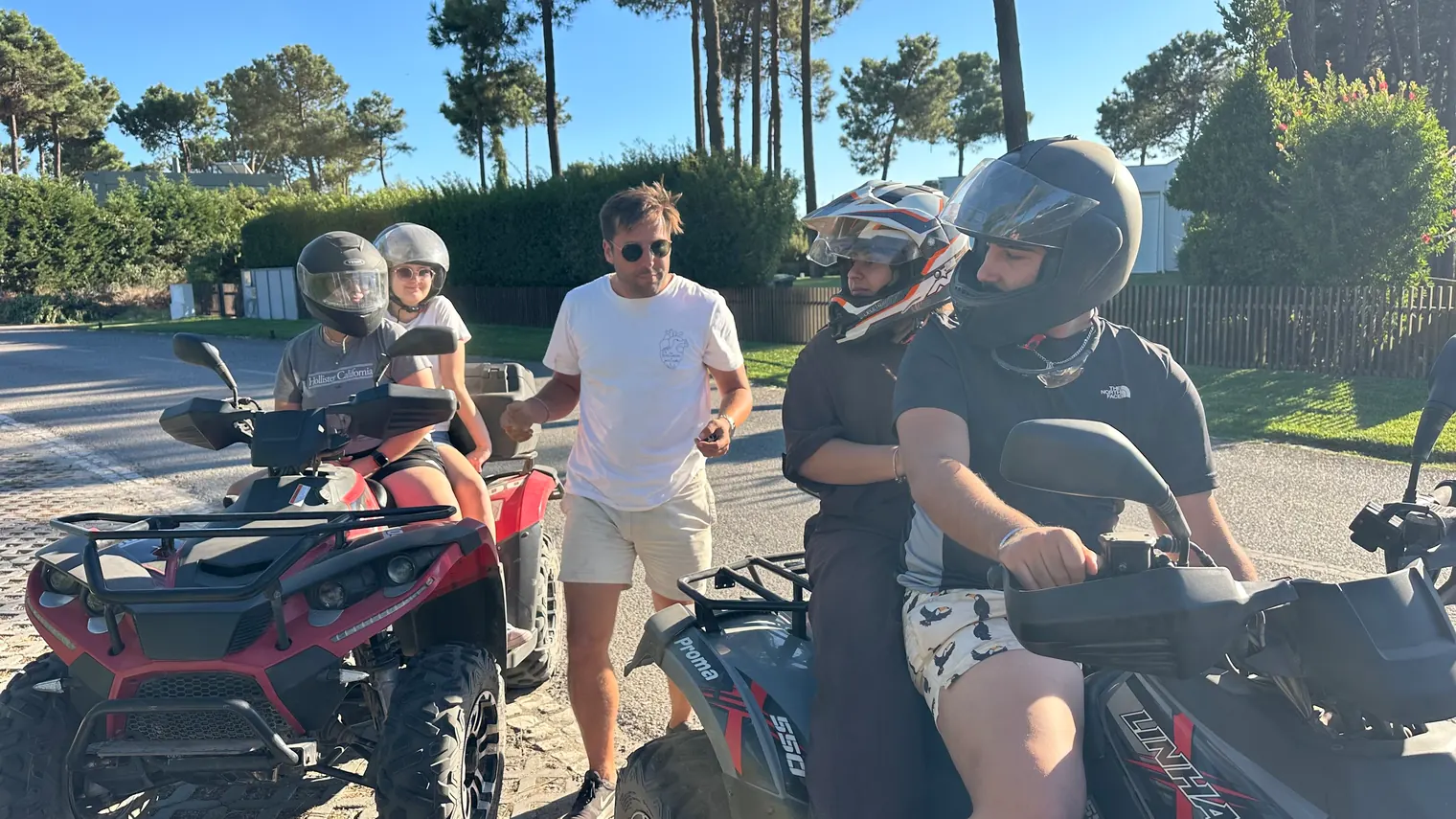 A group of people wearing helmets and standing or sitting on ATVs along a sunny tree-lined road.