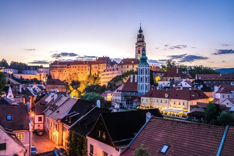 Twilight view of Český Krumlov with castle and tower, illuminated by warm lights. Red-roofed houses in the foreground under a purple sky.