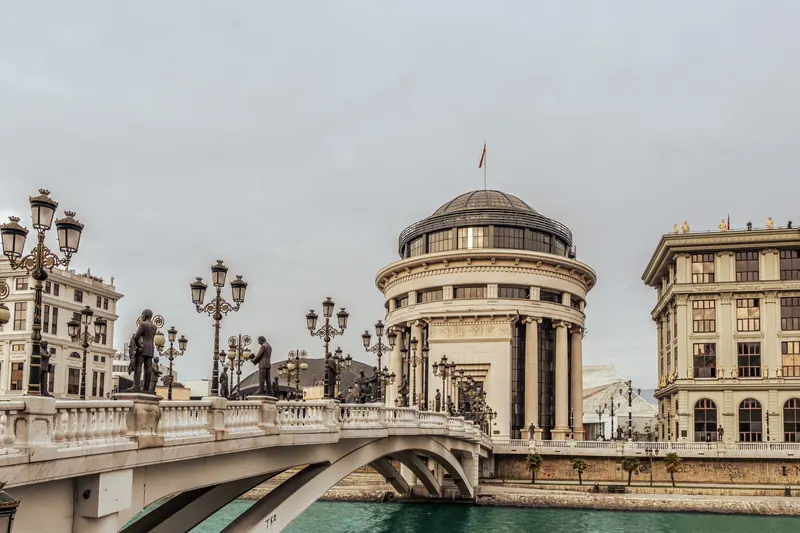 Ornate bridge with bronze statues and lampposts spans a river toward a domed neoclassical building under a gray sky.
