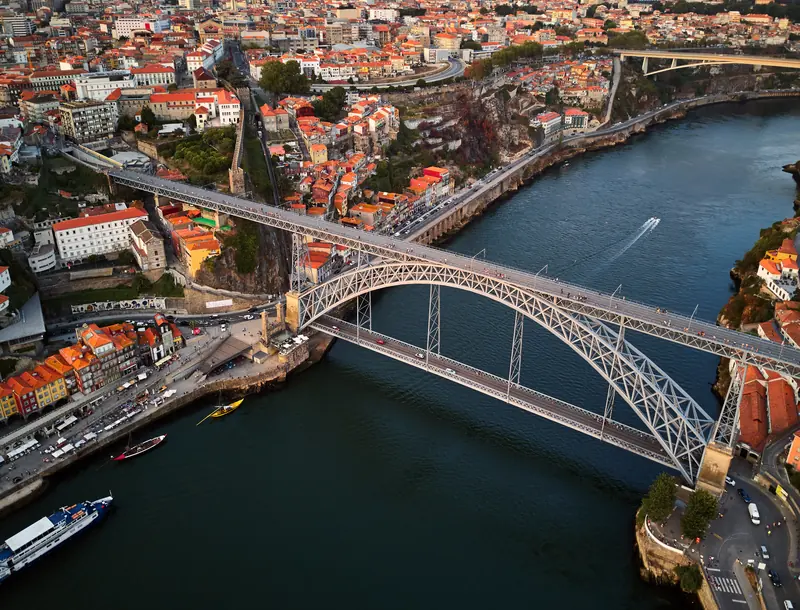 Aerial view of Porto’s Dom Luís I Bridge over the Douro River, with red-roofed city blocks, boats, and cliffs below.