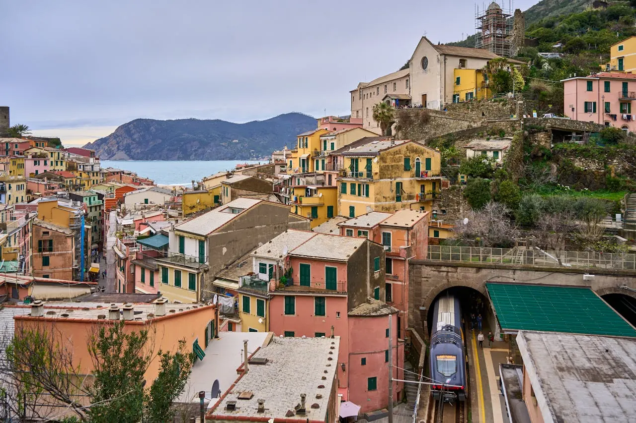 Coastal town with colorful buildings on a hillside, train emerging from a tunnel, sea and mountains in the background under a cloudy sky.