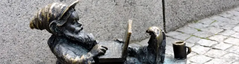 Laptop Dwarf. Bronze gnome statue reclines with a laptop and mug beside a stone wall on cobblestones.