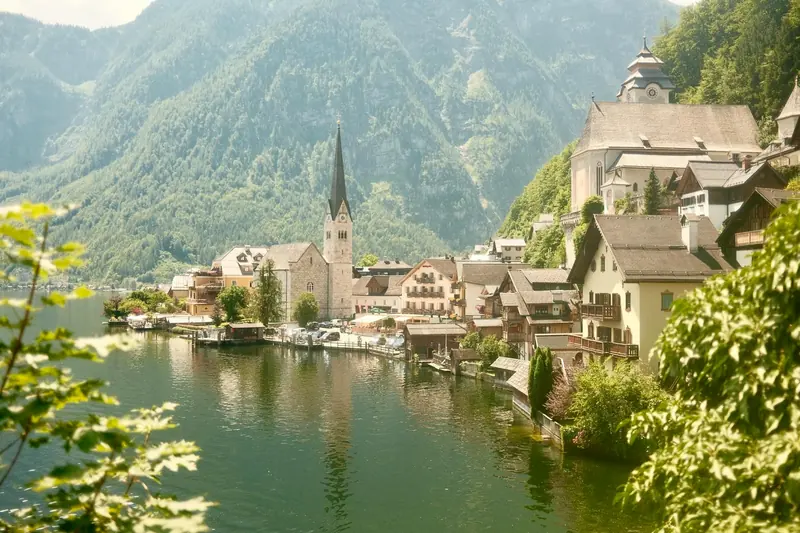 A serene lake scene with a boat sailing. A picturesque village with colorful houses. Majestic mountains and clear sky in the background.