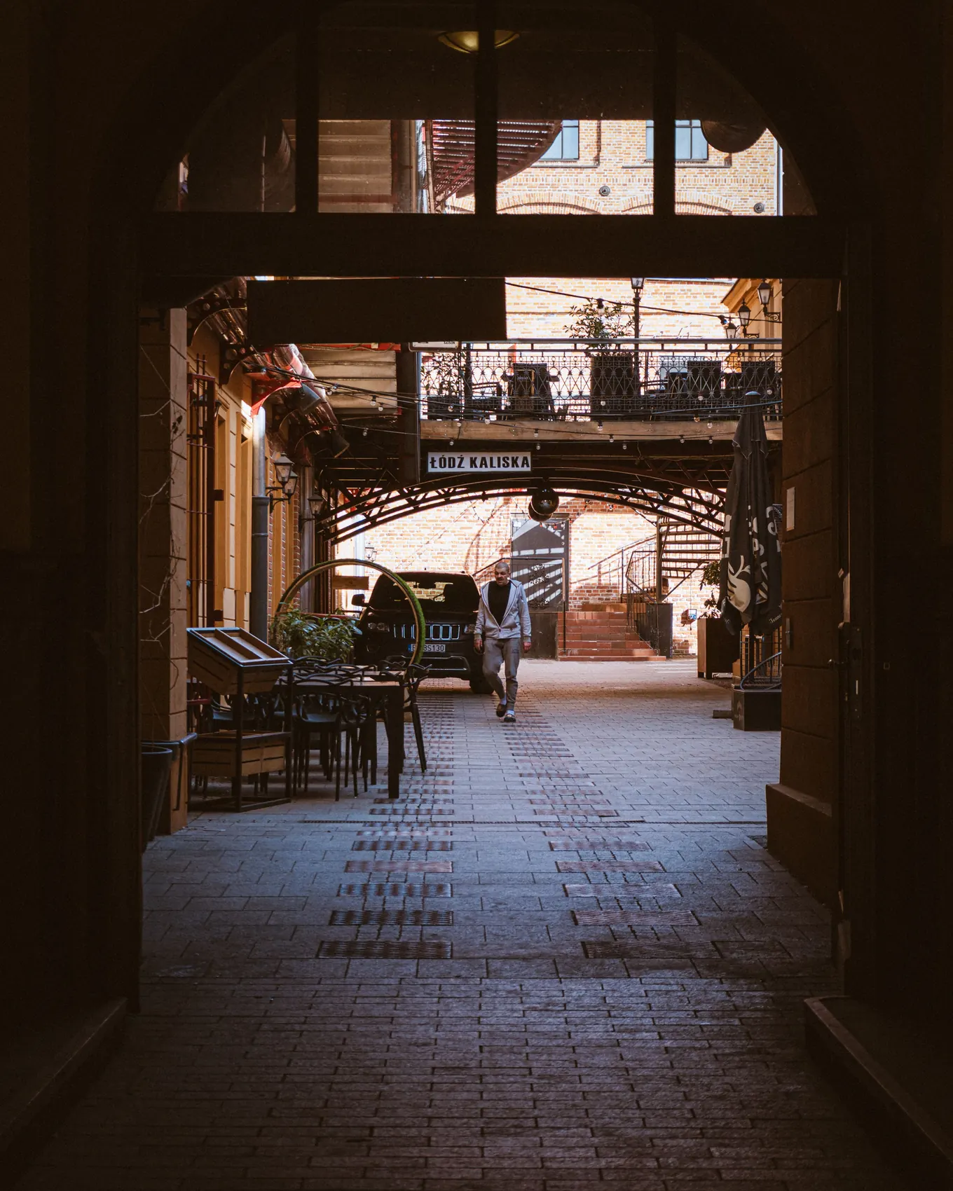 Man walks through a dim brick courtyard under a ŁÓDŹ KALISKA sign, with a parked SUV, tables, and warm sunlight.