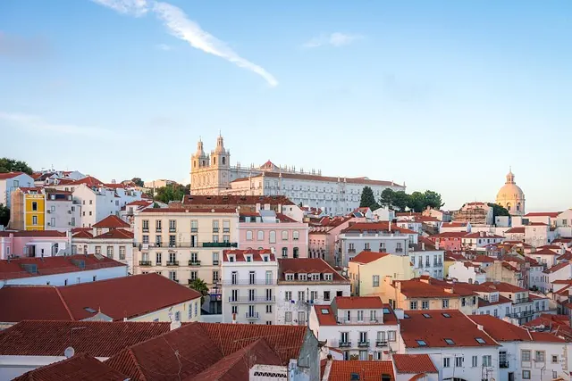 Rooftop view of Lisbon with red-tiled buildings under a clear blue sky. Historical church and dome in background. Calm and picturesque.