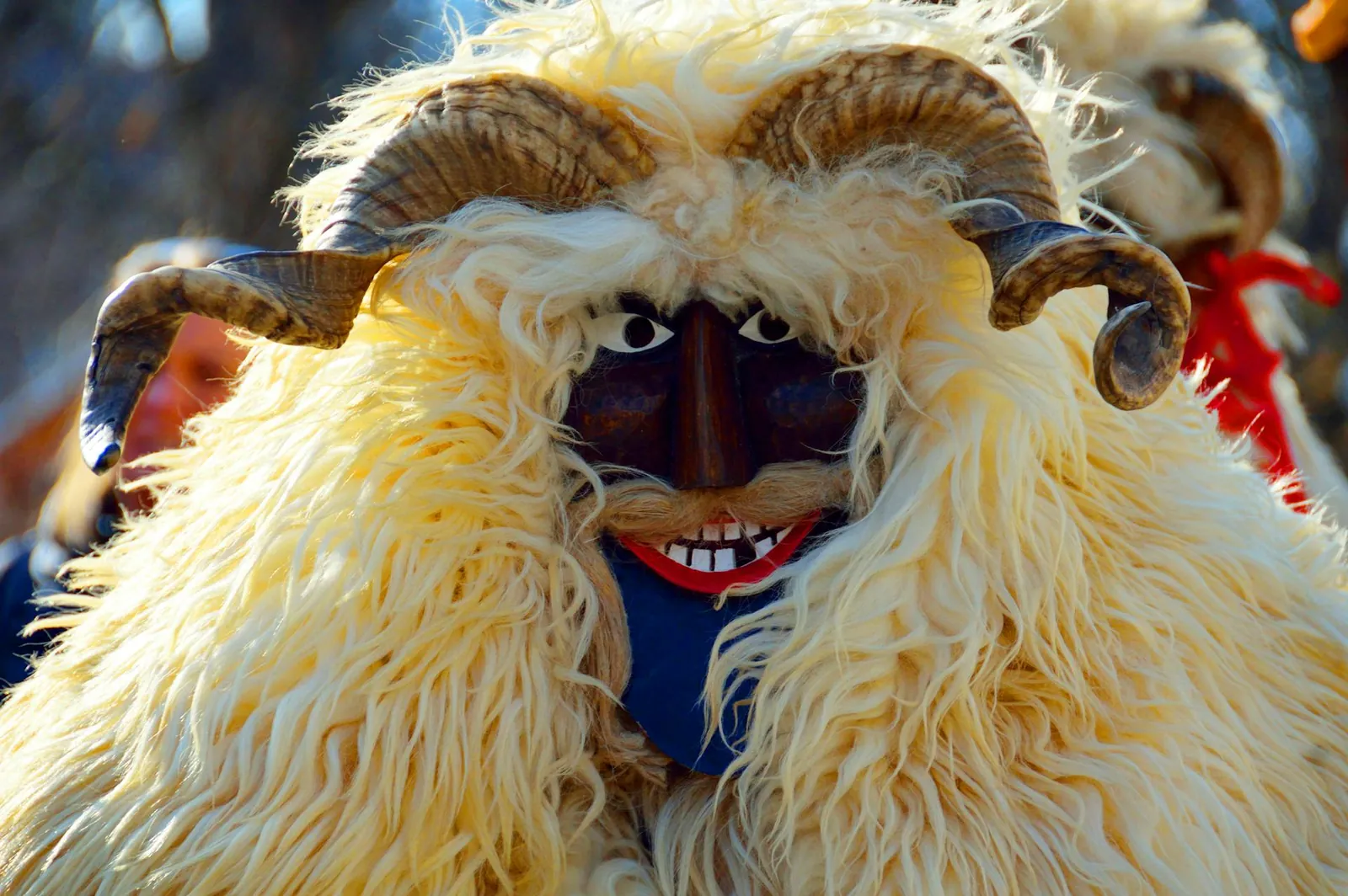 Person in a furry, horned costume with a painted mask and toothy grin. The background shows blurred people in colorful outfits. Bright and lively.