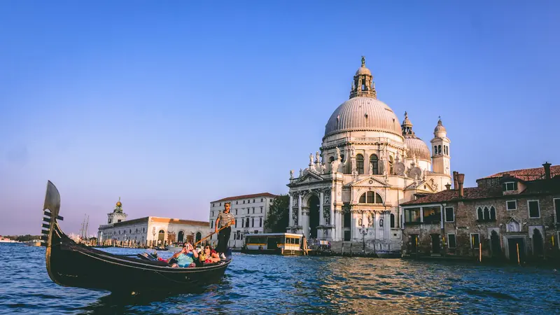 Gondola with passengers on Venice canal, passing a grand domed building at sunset. Clear blue sky and tranquil water set a serene mood.