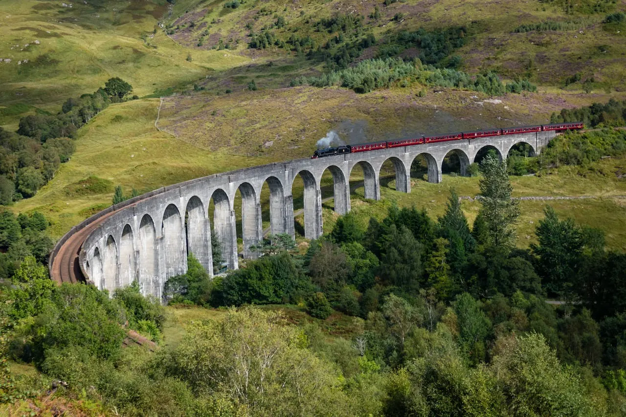 Train on a stone viaduct in lush green hills, emitting steam. Red carriages and vibrant landscape create a scenic, picturesque view.