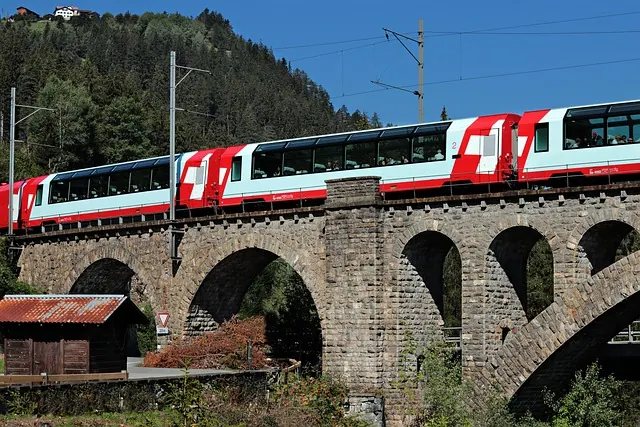 Train with red and white design crosses a stone viaduct in a forested area under a clear blue sky.