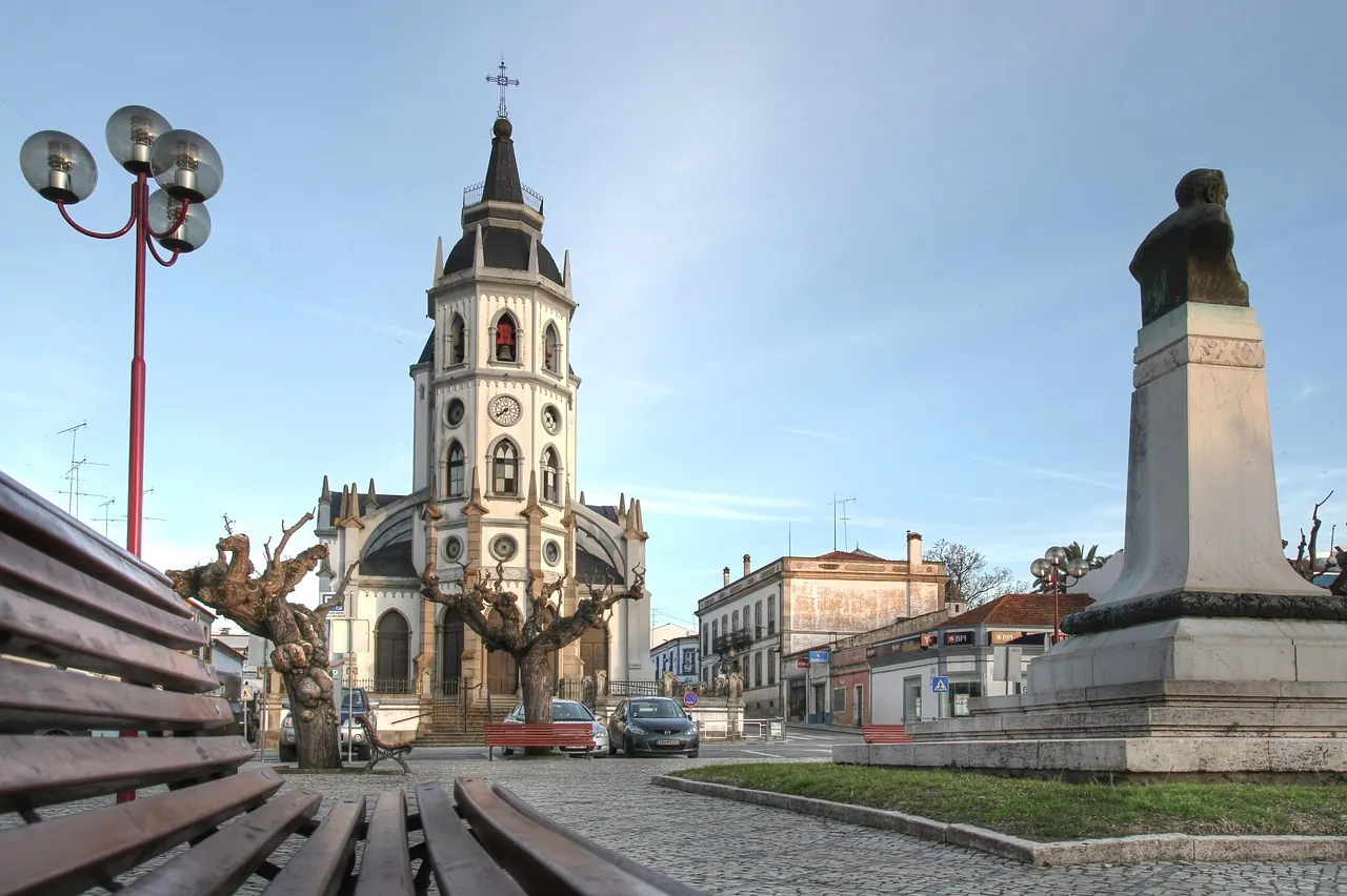 Church with a clock tower, bare trees, and a statue in a sunlit plaza. Cobblestone path and benches in the foreground. Calm atmosphere.
