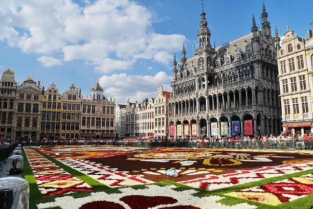 Colorful flower carpet in Brussels' Grand Place, surrounded by ornate buildings and a clear blue sky, with a crowd of onlookers.