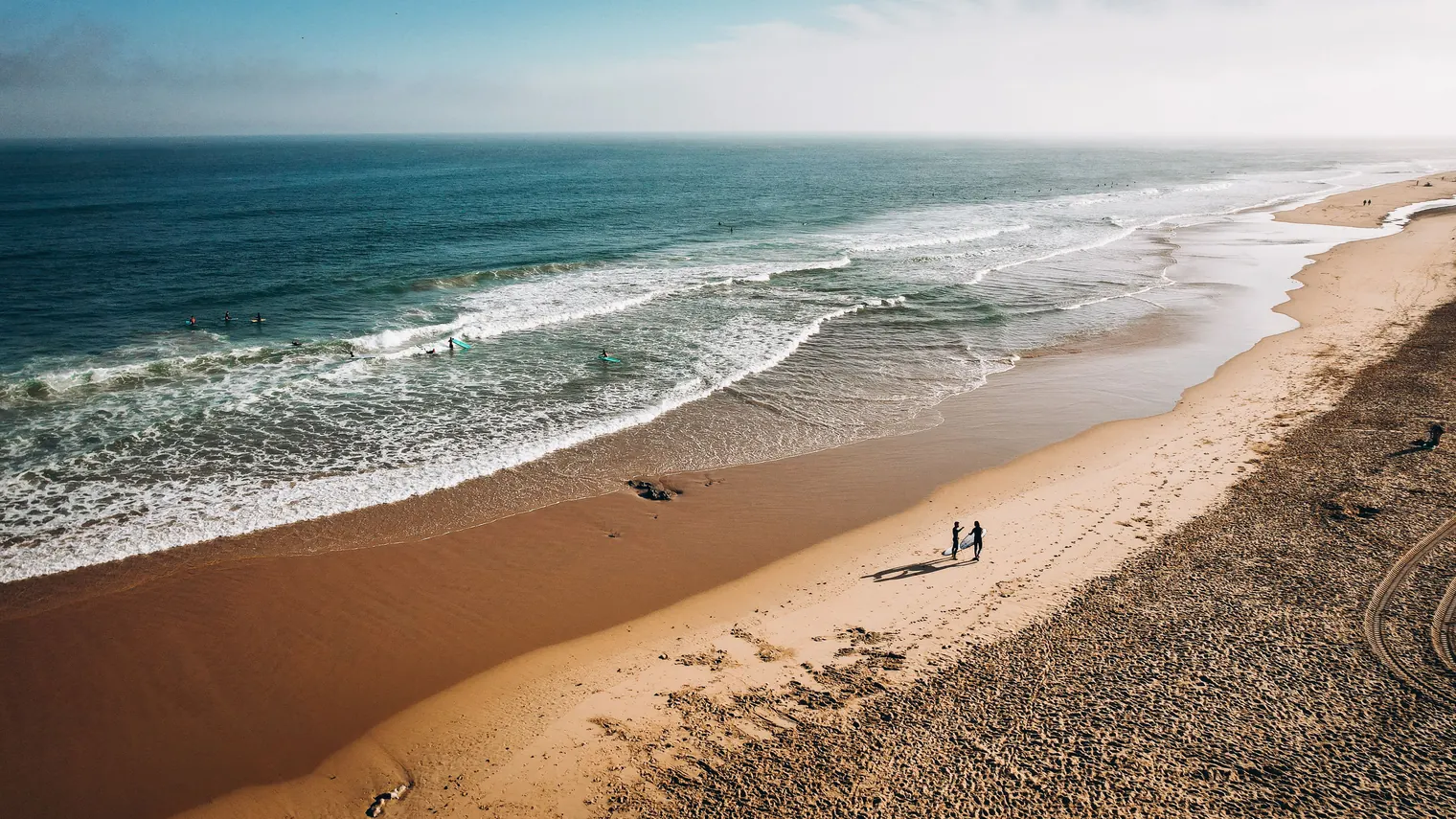 Aerial view of Costa da Caparica beach with golden sand, Atlantic waves, surfers, and people walking along the shoreline.