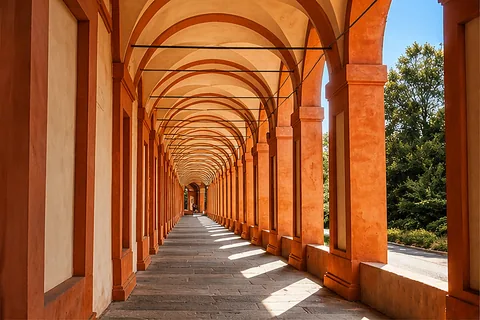 A long covered walkway with terracotta-colored columns and vaulted arches, sunlight casting geometric shadows across the stone floor, and green trees visible through the open side.